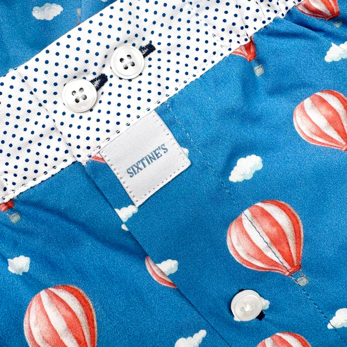 Close-up of a children's shirt with a blue body printed with red-and-white hot air balloons and small white clouds, plus a white polka-dot collar and white buttons. A fabric label reads SIXTINE'S.