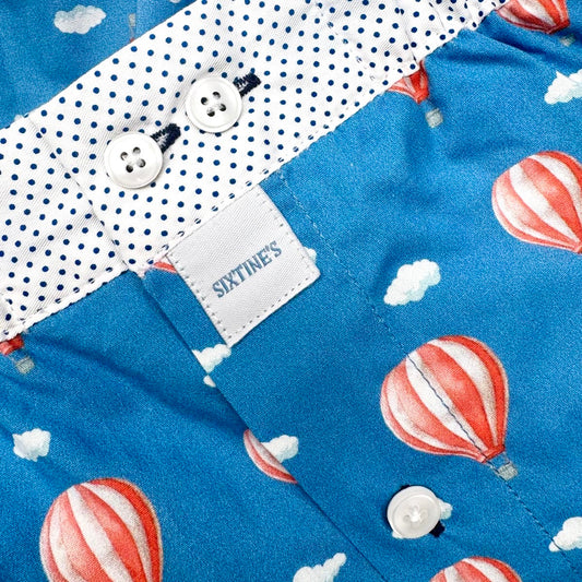 Close-up of a children's shirt with a blue body printed with red-and-white hot air balloons and small white clouds, plus a white polka-dot collar and white buttons. A fabric label reads SIXTINE'S.