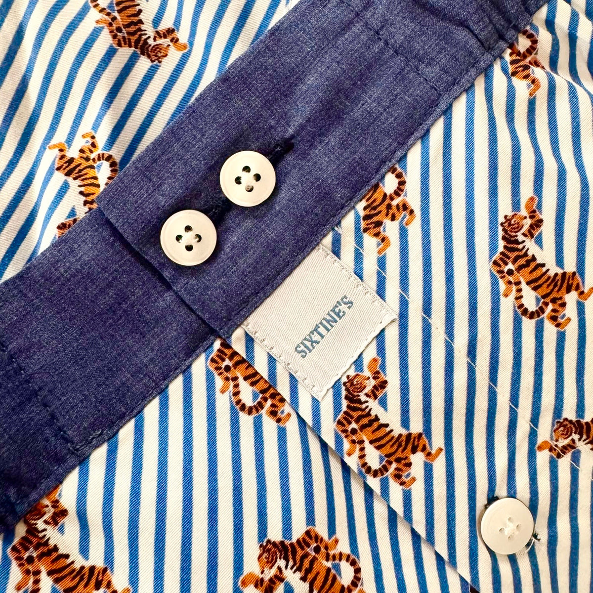 Close-up of a children's shirt or pajama top showing a blue and white striped fabric with orange and black tiger print, a dark blue collar, and white buttons. A fabric label reads SIXTINES.